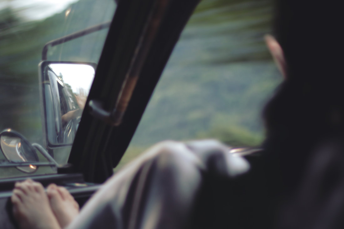 A photograph of the view from inside a moving van, 2018. The camera is pointed at a passenger in the front seat who has their bare feet on the dashboard. The mirrors that can be seen through the windshield shows the passenger has their hand stuck outside the window feeling the wind. The cool green mountainous terrain outside is a blur from the speed.