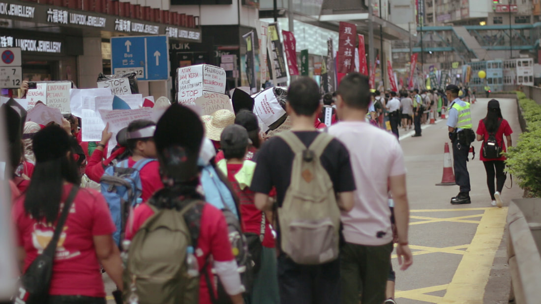 A photograph of a May Day street procession in Hong Kong, 2019. Demonstrators have backs facing the camera, in the foreground is a contingent of migrant domestic workers, many of whom are wearing red t-shirts and carrying bags, holding handwritten protest signs big and small, some even on their heads, in Indonesian. Some signs read 