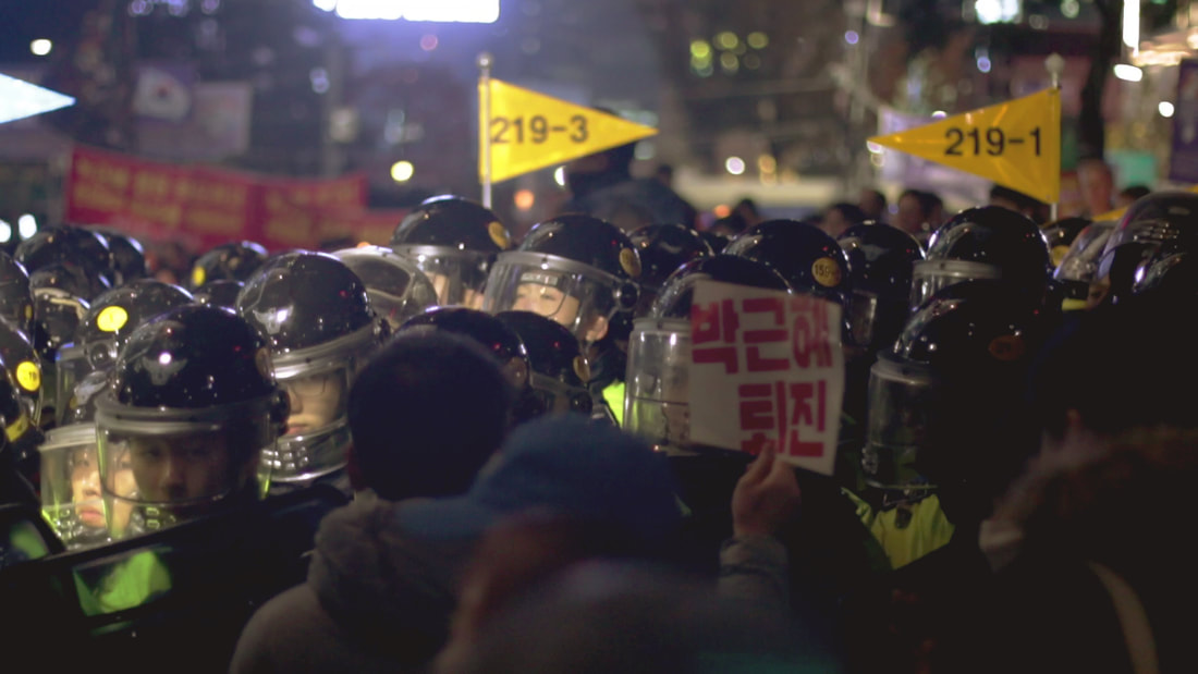 A photograph of an anti-government demonstration in Seoul, 2016. Protestors in the foreground are pushed up face-to-face against a tightly packed contingent of police in anti-riot gear. In the foreground a protestor is holding up a printed sign in Korean that reads 